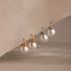 Two pairs of Pearl Dome Drop Earrings, each featuring lustrous pearls—one with gold hooks and one with silver—are displayed on a textured surface. Perfect for any occasion.