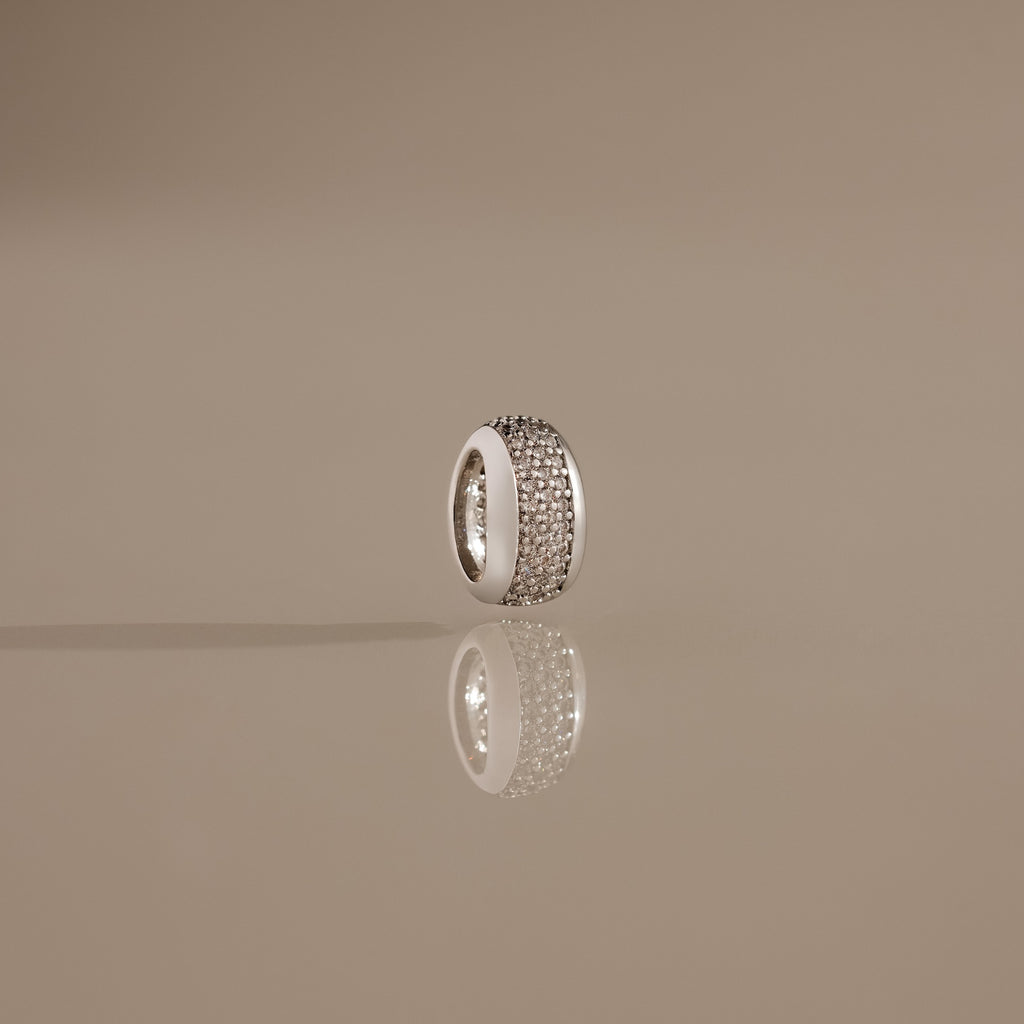 The Bold Pave Dome Charm in Silver, adorned with rows of sparkling stones and standing upright on a reflective beige surface, showcases timeless elegance in every detail.
