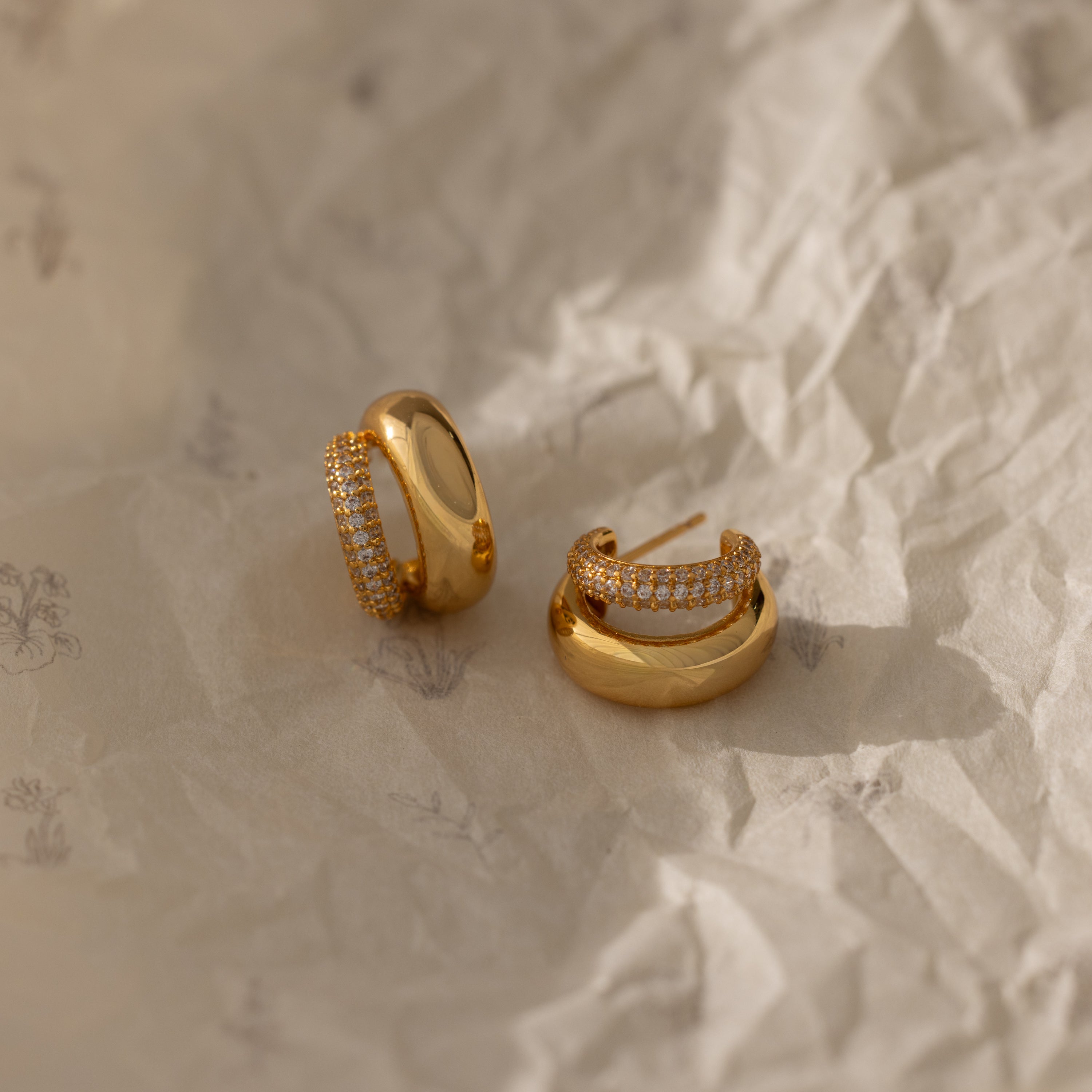 The Duo Pave Dome Studs in 18K Gold rest on crumpled, light-colored paper—a stunning pair of earrings perfect for everyday jewelry lovers.