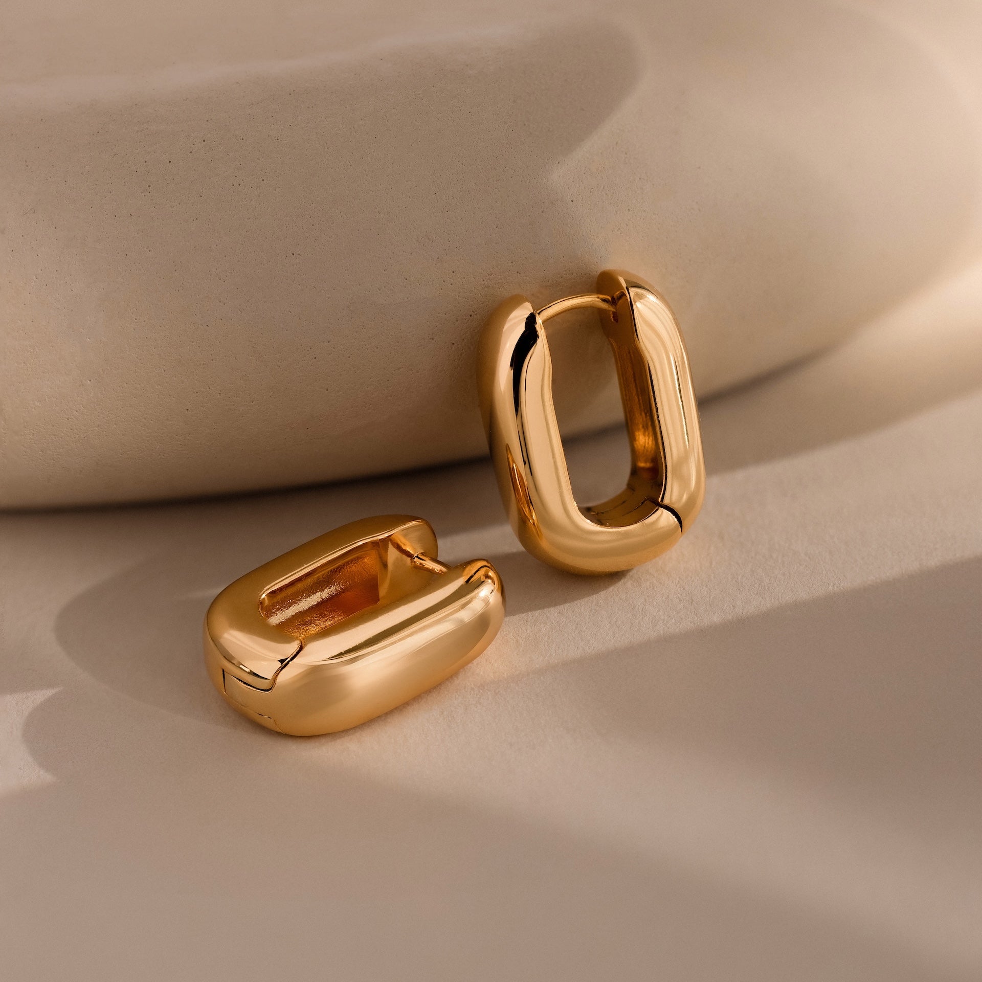The Bold Link Hoops in 18K Gold are chunky, U-shaped earrings shown on a beige, softly lit surface—perfect modern statement pieces.