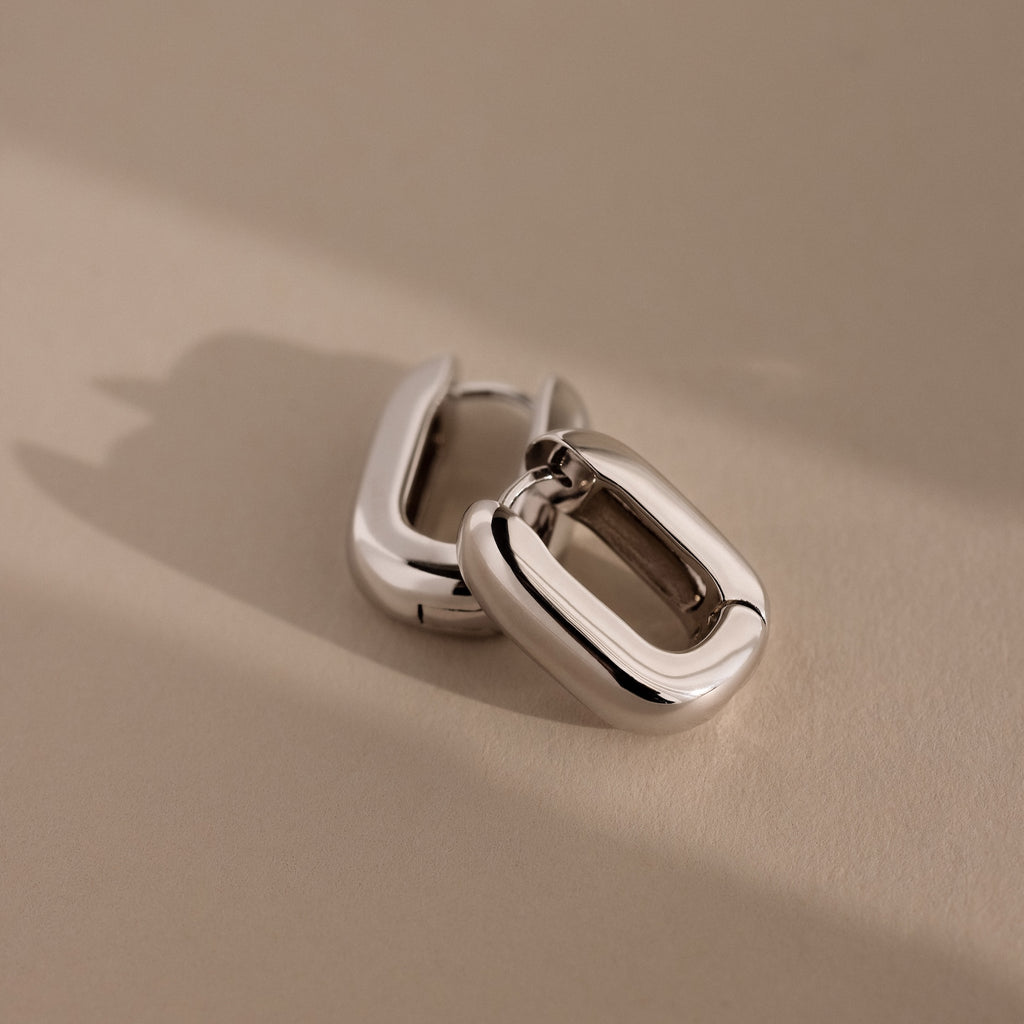 Two chunky Bold Link Hoops—sleek, rectangular silver earrings—rest on a beige surface with soft shadows, embodying modern jewelry style.