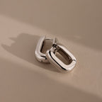Two chunky Bold Link Hoops—sleek, rectangular silver earrings—rest on a beige surface with soft shadows, embodying modern jewelry style.