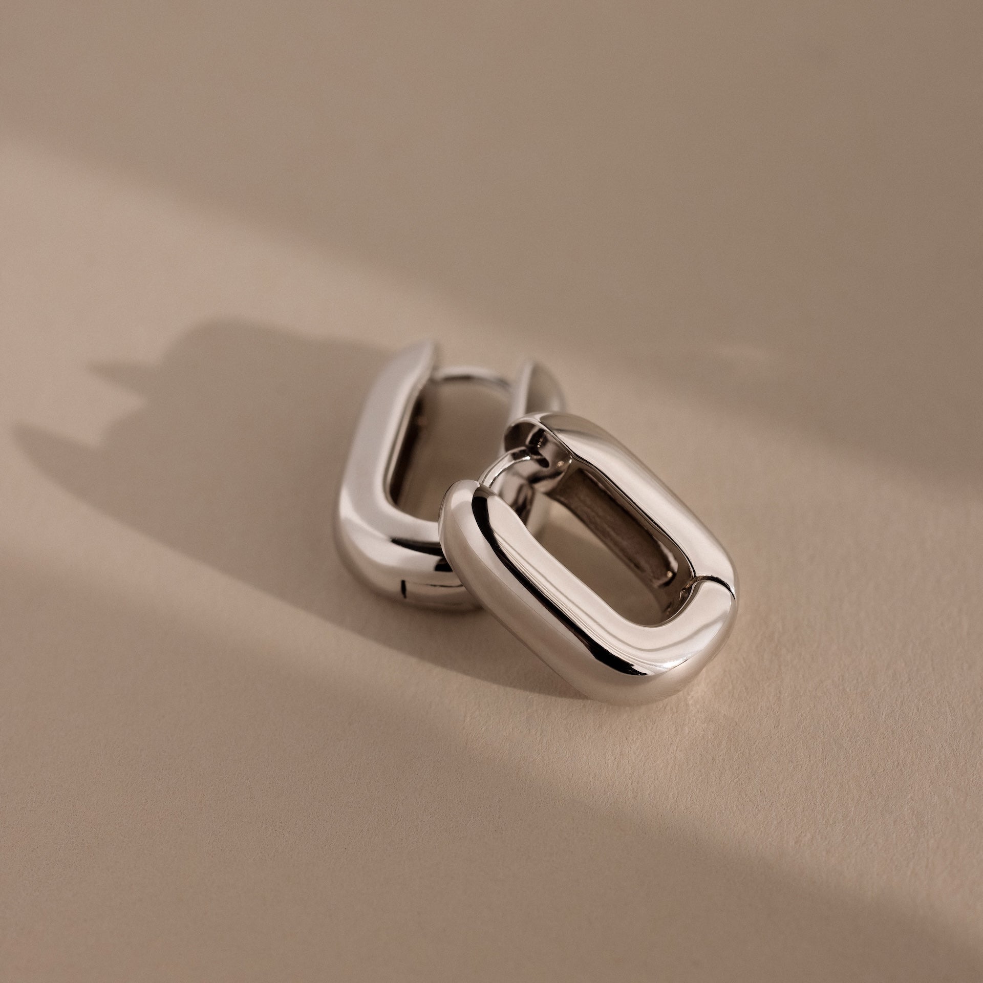 Two chunky Bold Link Hoops—sleek, rectangular silver earrings—rest on a beige surface with soft shadows, embodying modern jewelry style.