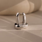 The Bold Link Hoops in White Gold, from our signature collection, stand upright on a beige surface with soft shadows—perfect for fans of modern jewelry.