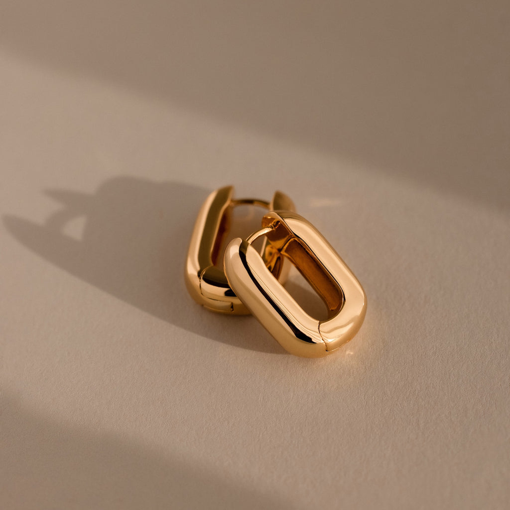 The Bold Link Hoops in 18K Gold, featuring a chunky rectangular design, rest on a neutral surface with soft shadows—perfect for adding modern style to any look.