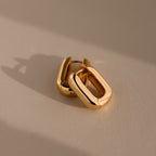 The Bold Link Hoops in 18K Gold, featuring a chunky rectangular design, rest on a neutral surface with soft shadows—perfect for adding modern style to any look.