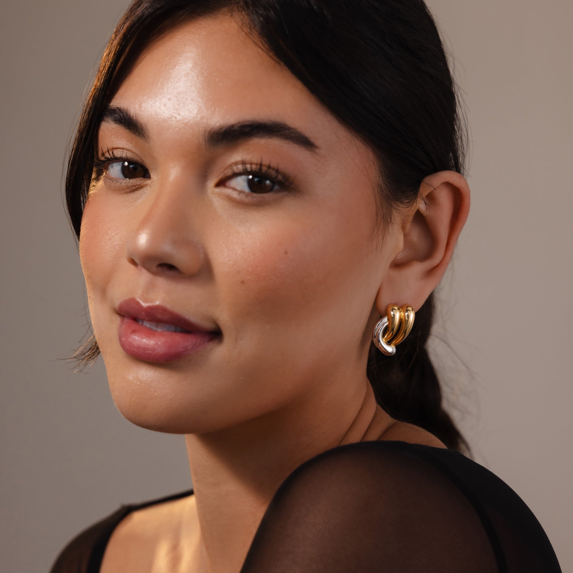 Woman with long dark hair smiles softly at the camera, wearing a black sheer top and Mixed Metal Raindrop Studs in gold and silver.