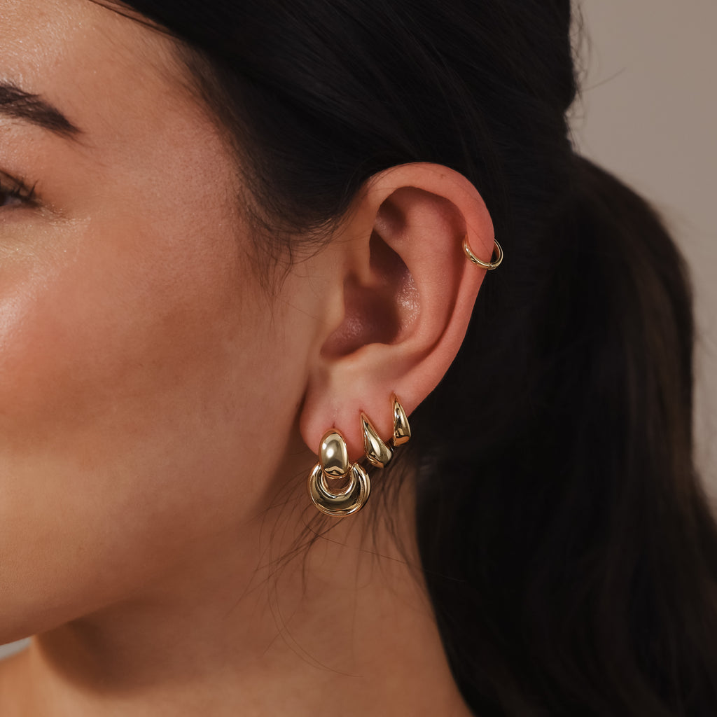 Close-up of a woman’s ear adorned with multiple gold earrings, including the Eclipse Dome Studs in 18K Gold and a small hoop in the upper cartilage, showcasing a modern jewelry style.