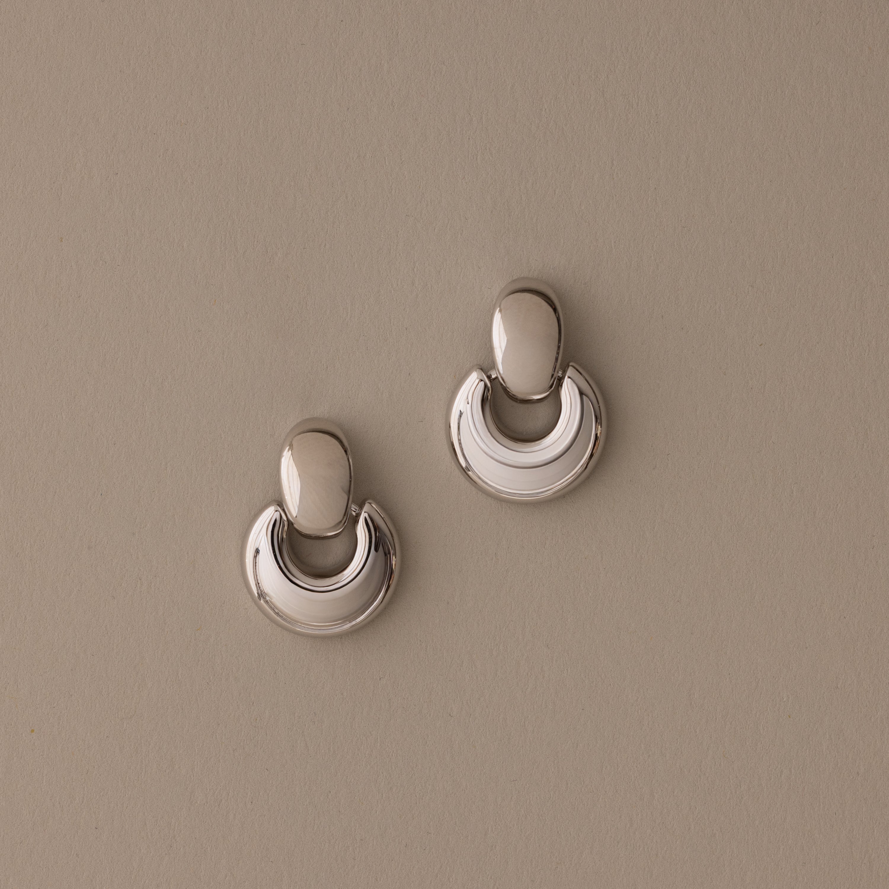 A pair of Eclipse Dome Studs in White Gold, featuring a shiny, crescent-shaped sculptural design, rests on a plain beige background.