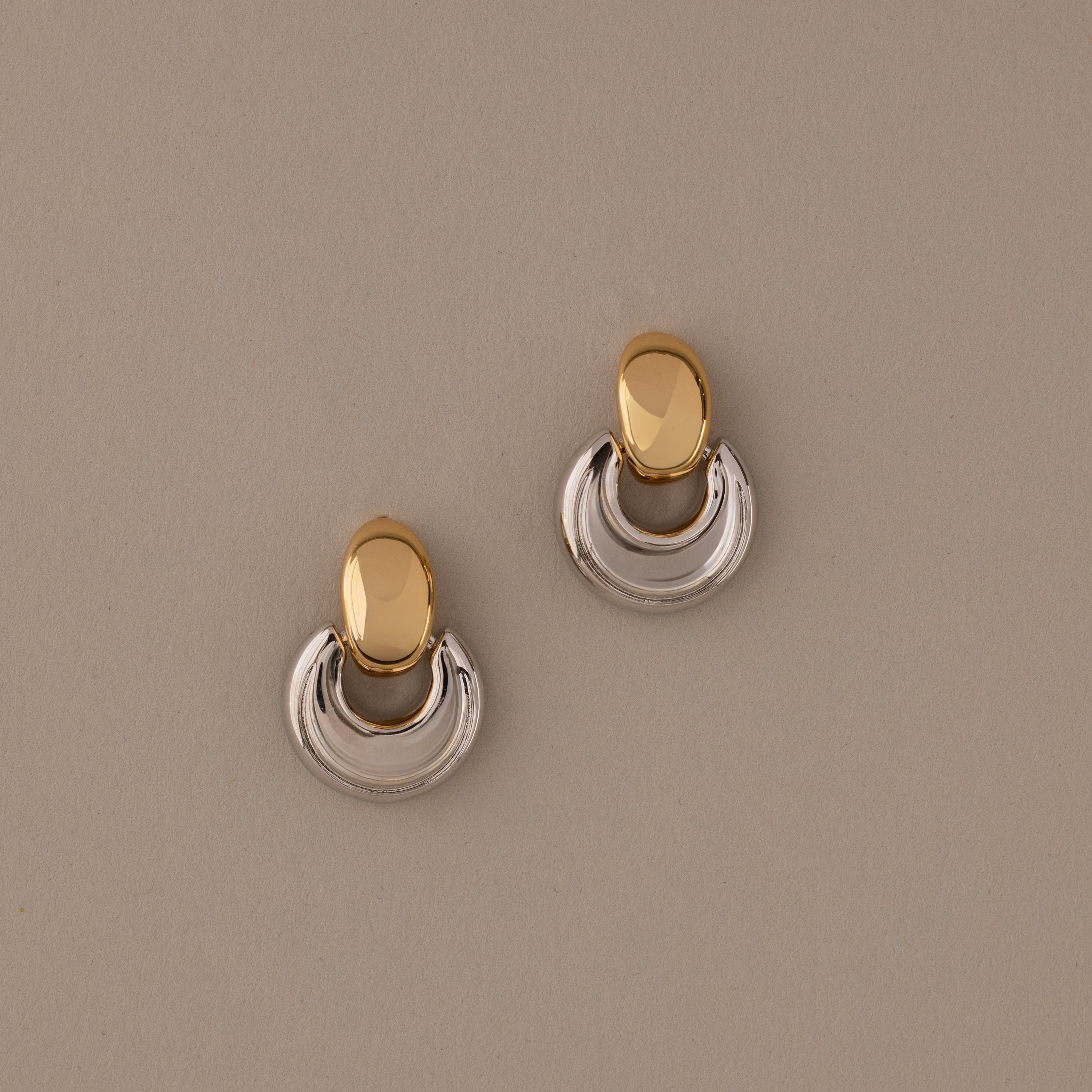 The Eclipse Dome Studs in Mixed Metal feature oval tops and curved half-moon bottoms, highlighting sculptural design against a beige background.