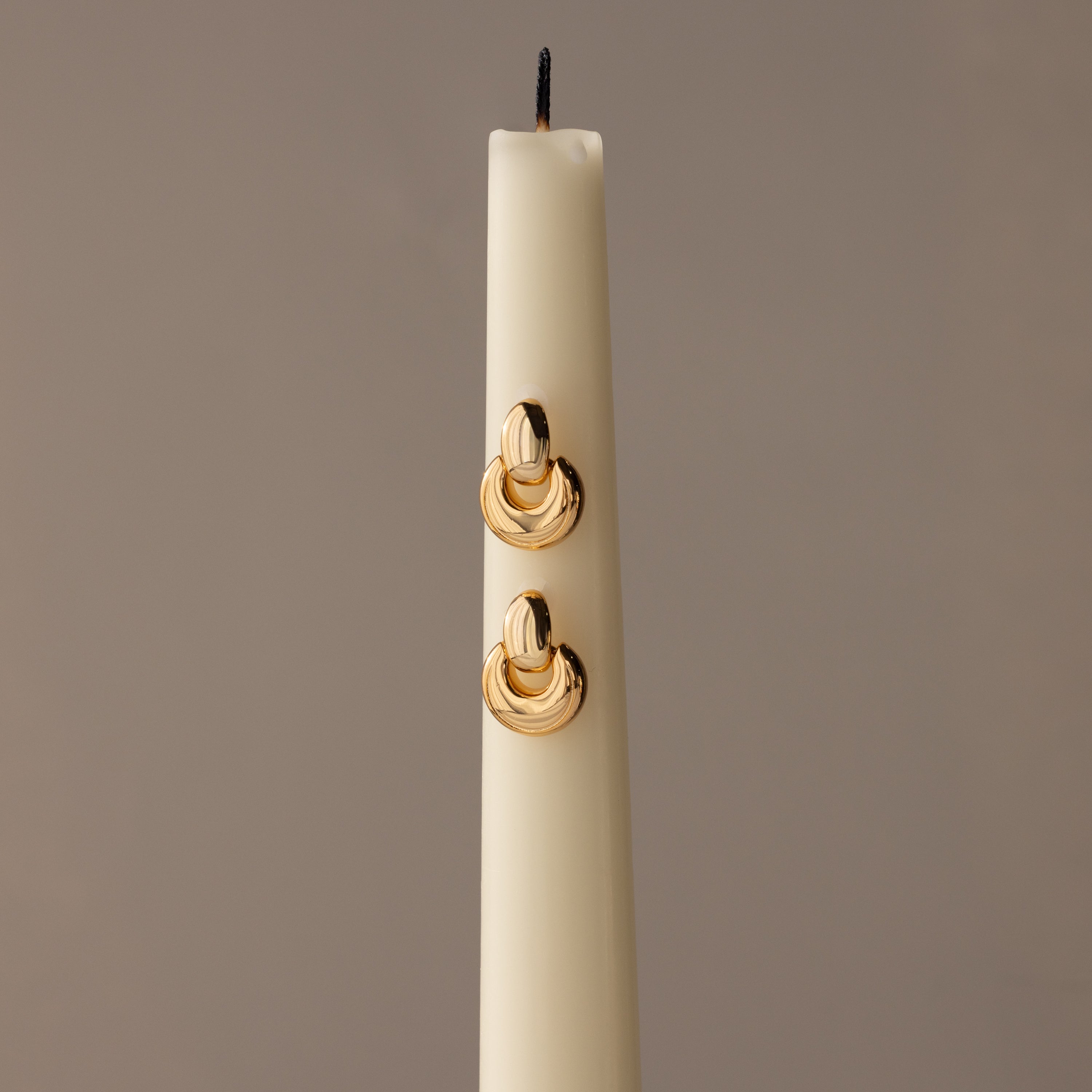 A white candle displays sculptural earrings and Eclipse Dome Studs in 18K gold, set against a neutral background for a modern jewelry showcase.