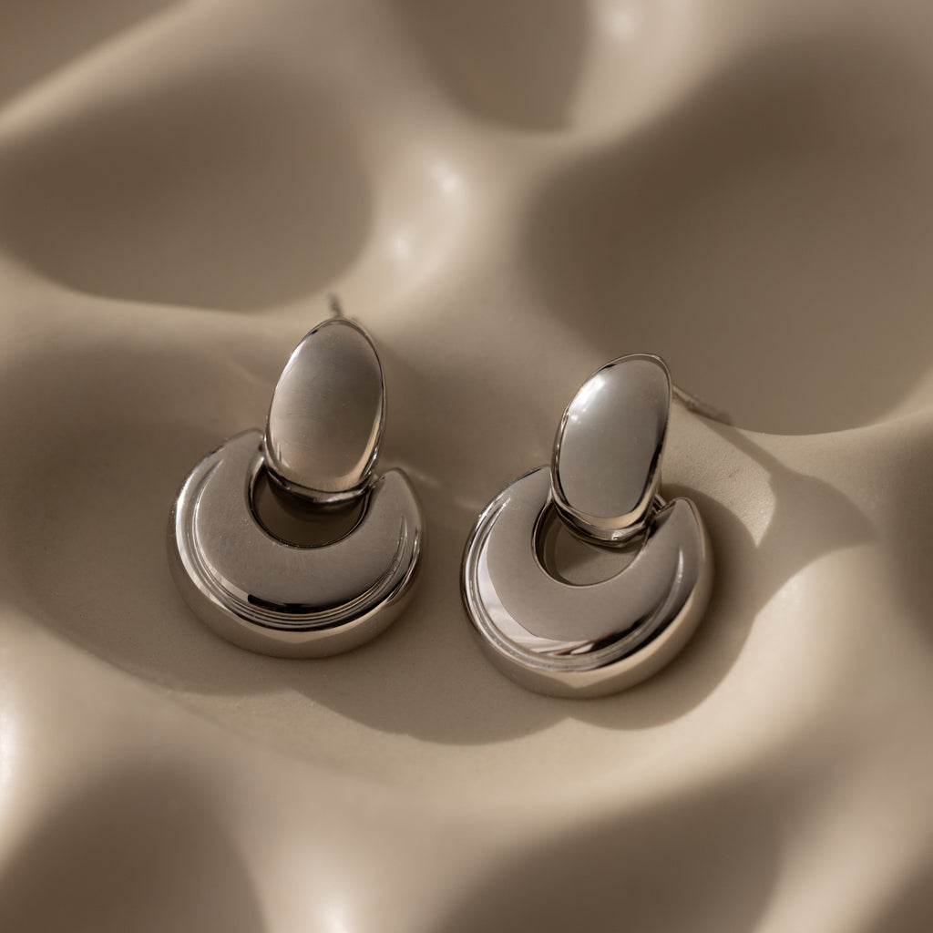 Eclipse Dome Studs in White Gold, sculptural crescent-shaped earrings, rest on a softly textured beige surface, reflecting light—modern jewelry with subtle elegance.