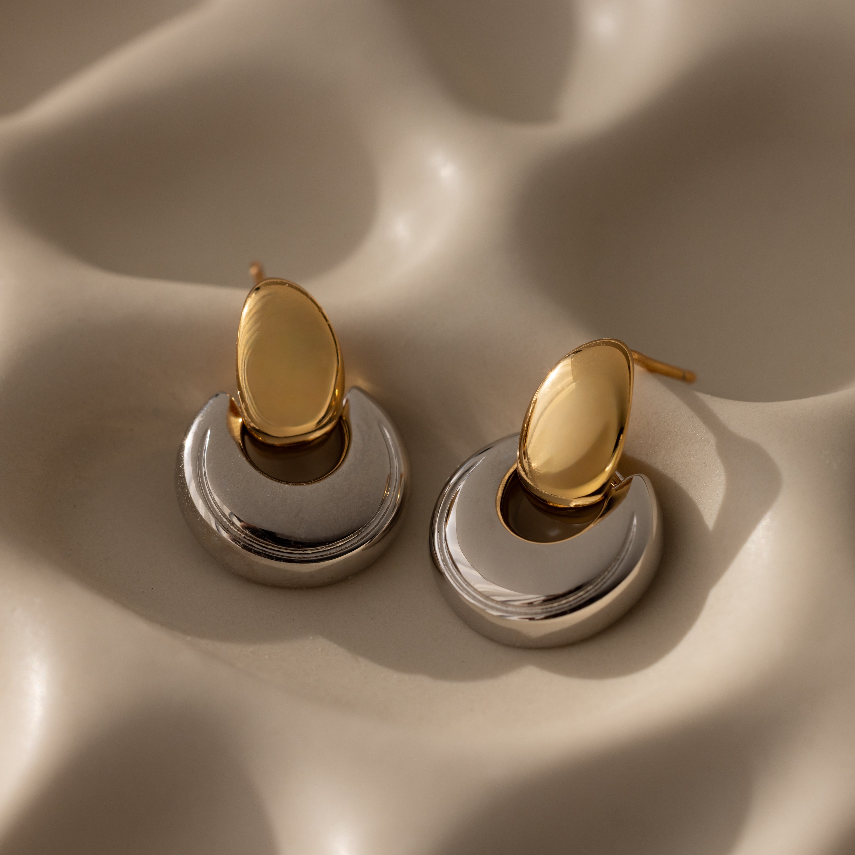 The Eclipse Dome Studs in Mixed Metal, featuring gold and silver crescent shapes, rest on a cream wavy textured surface, highlighting the elegance of modern jewelry design.