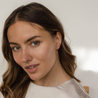Woman with wavy brown hair and a light-colored top smiles softly at the camera, showcasing the Dainty Pearl Studs in 18K Gold.