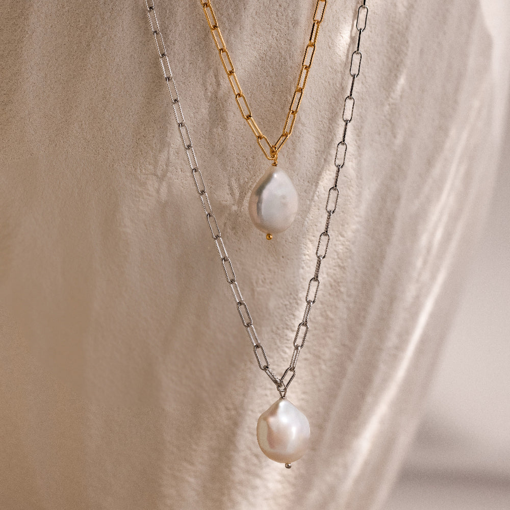 Two pearl pendant chain necklaces in gold and silver on a beige stone background