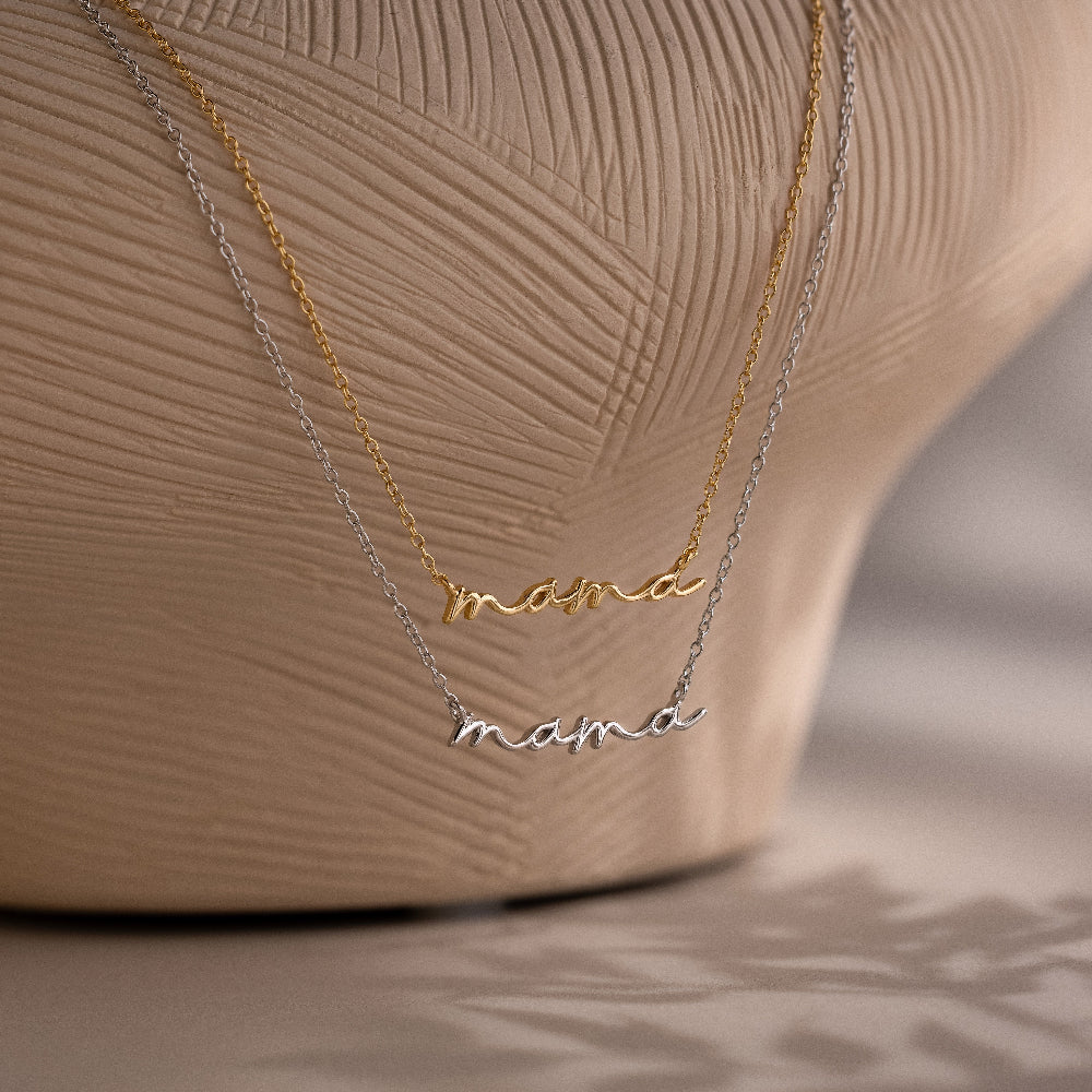 Two 'mama' pendant necklaces in gold and silver on a textured beige background