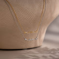 Two 'mama' pendant necklaces in gold and silver on a textured beige background