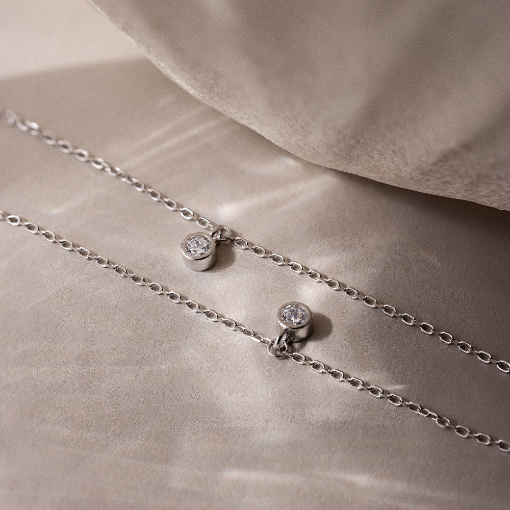 Two Diamond Station Necklaces in Silver, featuring small round diamond pendants, rest on a soft beige surface—embodying modern elegance for a timeless accessory to enhance any look.