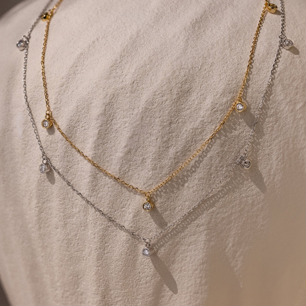 The Diamond Station Necklace in Silver and gold, adorned with small round gems, captures modern elegance as it rests on textured fabric.