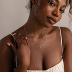 A woman in a cream dress touches the Diamond Station Necklace in Silver, highlighting its modern elegance against a neutral background.