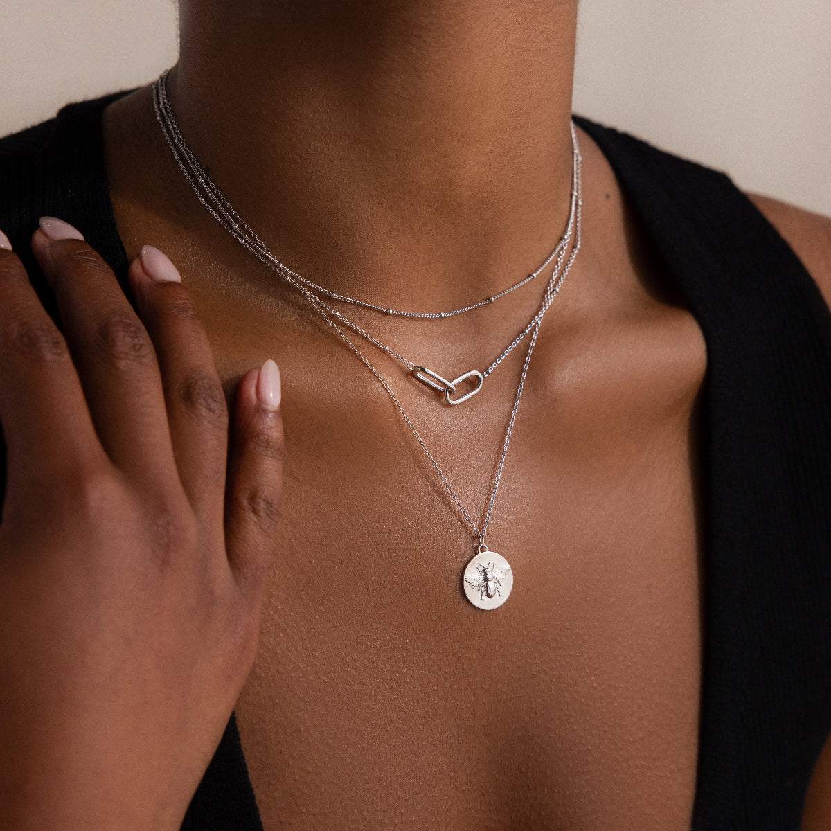 A woman wears the Satellite Necklace in Silver, layered with other silver necklaces, paired with a black sleeveless top and her hand resting on her chest.
