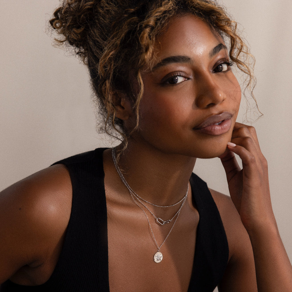 A woman with curly hair and a black top smiles softly, touching her face while elegantly showcasing layered necklaces, including a simple chain and the Satellite Necklace in Silver—perfect for layering.