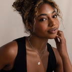 A woman with curly hair and a black top smiles softly, touching her face while elegantly showcasing layered necklaces, including a simple chain and the Satellite Necklace in Silver—perfect for layering.