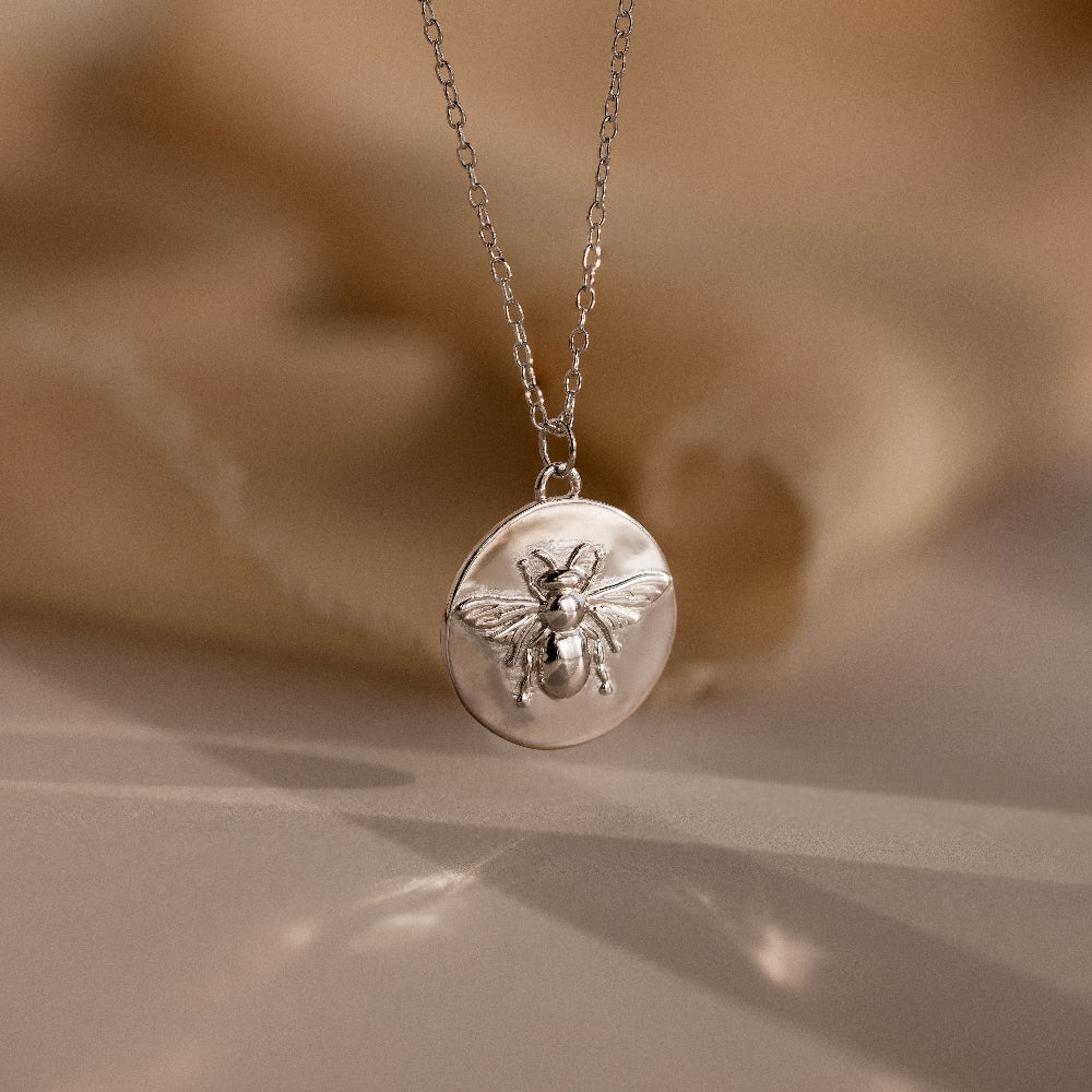 The Bee Coin Necklace in Silver features a round pendant with a raised bee design and is perfect for layering on any occasion. Displayed against a soft beige background.