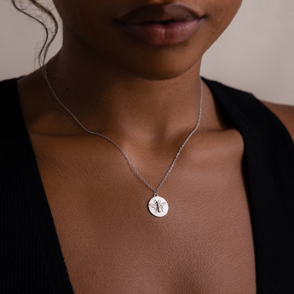 A woman wears a black top and the Bee Coin Necklace in Silver, featuring a round pendant with an insect design.
