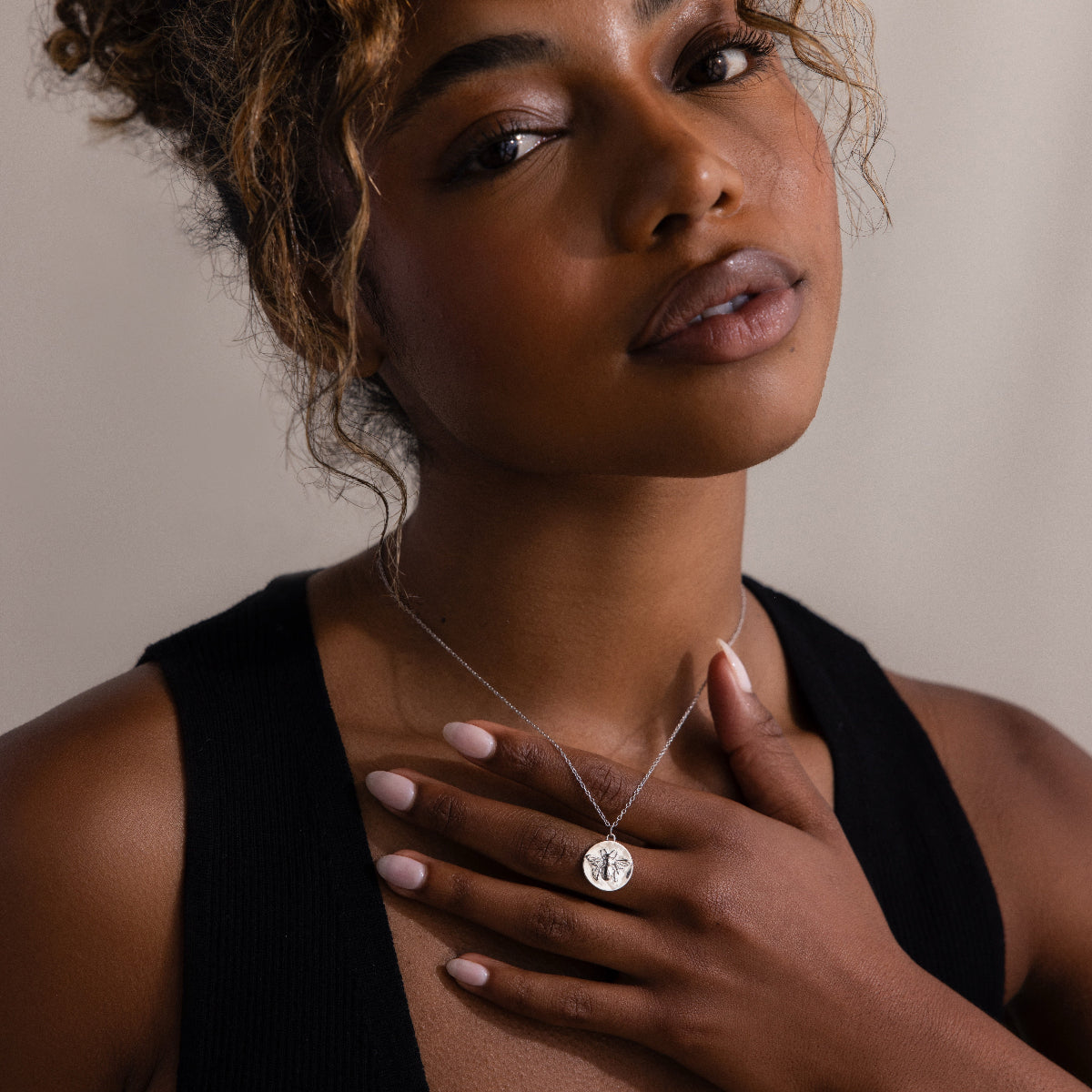 Woman in a black top touches the Bee Coin Necklace in Silver, gazing softly at the camera against a neutral background.
