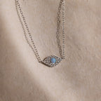 The Opal Eye Necklace in Silver features a blue stone and small clear crystals, creating a striking lucky talisman, showcased on a beige surface.