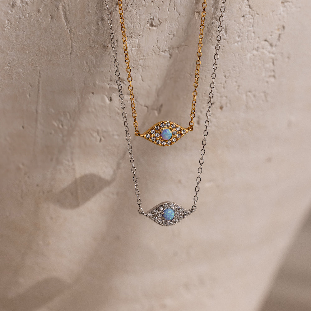 The Opal Eye Necklace in silver and gold features a blue stone center in an eye-shaped pendant—an elegant talisman, perfect for adding luck and style on a beige background.