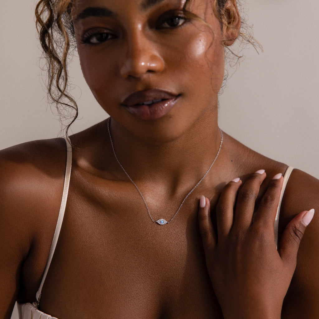 Woman wearing the Opal Eye Necklace in Silver, featuring a blue opal pendant, touches her shoulder under soft lighting—an elegant talisman for any occasion.