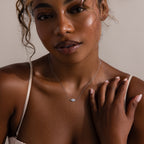 Woman wearing the Opal Eye Necklace in Silver, featuring a blue opal pendant, touches her shoulder under soft lighting—an elegant talisman for any occasion.