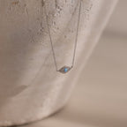 The Opal Eye Necklace in Silver features a delicate blue gemstone pendant, shown on a textured, light background—an elegant piece perfect as your lucky talisman.
