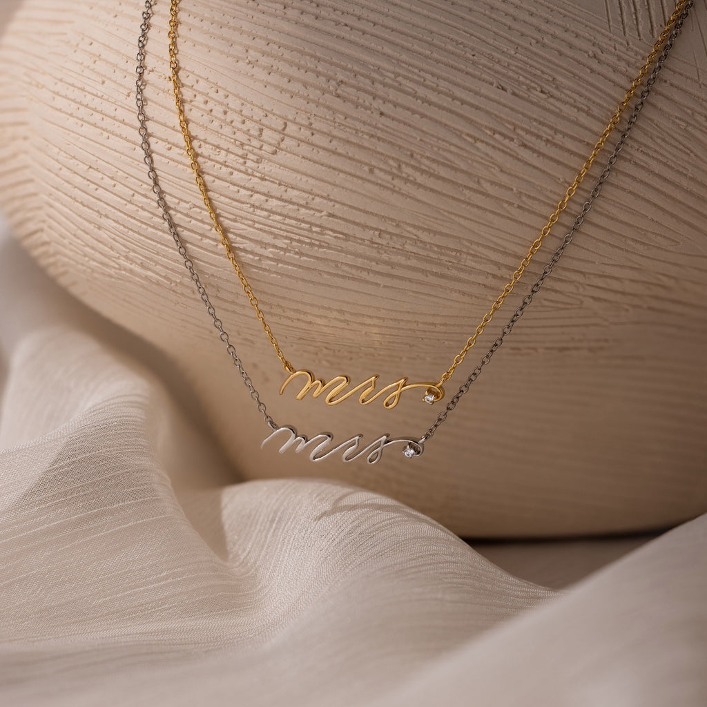 The Mrs. Necklace in silver and gold is displayed on a textured neutral background—an elegant and thoughtful wedding gift for any bride-to-be.