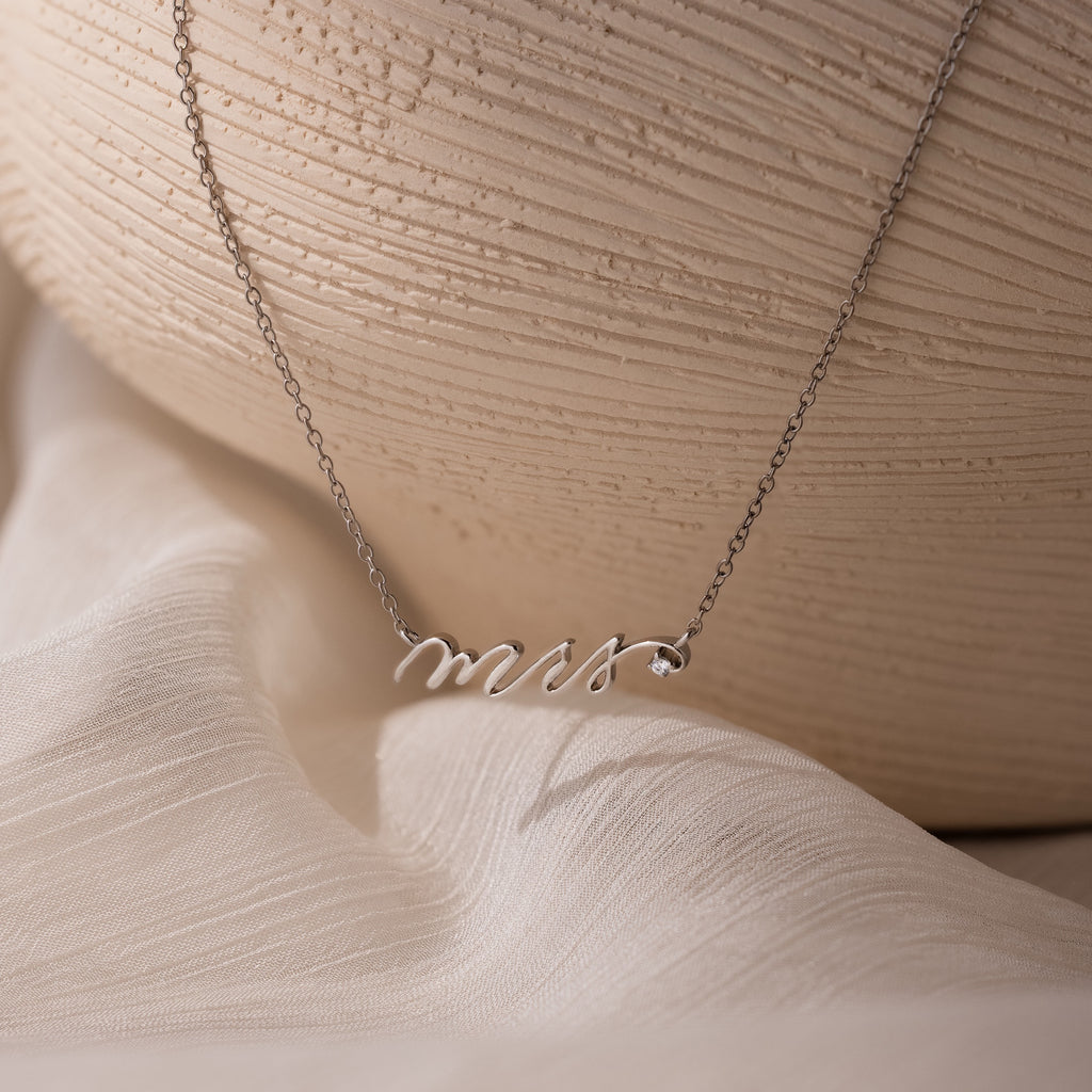 The Mrs. Necklace in Silver with a Mrs pendant is displayed on soft white fabric next to a ceramic vase—an ideal, thoughtful wedding gift for the bride-to-be.
