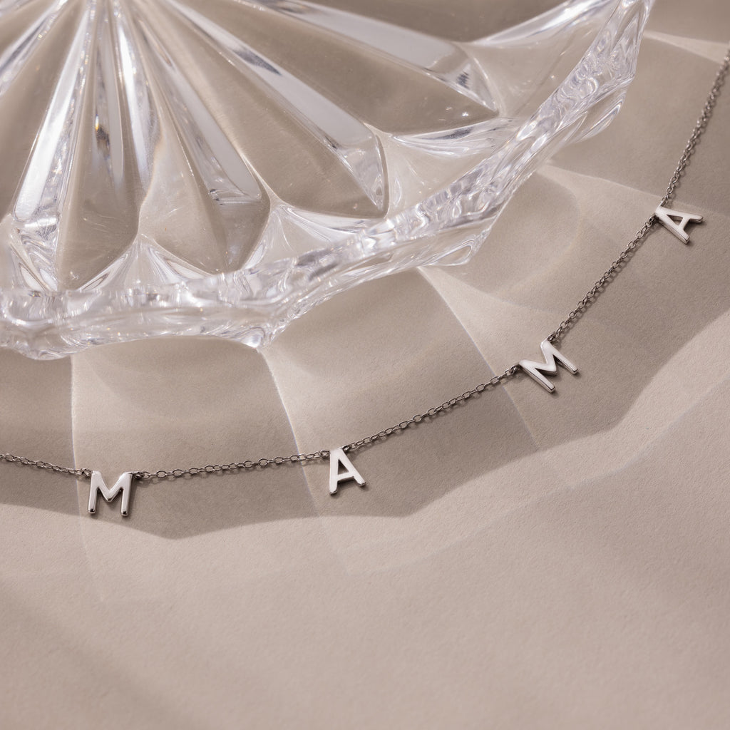 The Mama Letter Necklace in Silver spells “MAMA” and rests beside a clear glass dish on a beige surface—a beautiful tribute to motherhood and the perfect gift for any mom.
