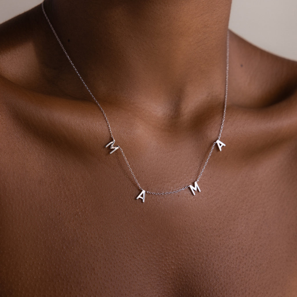 The Mama Letter Necklace in Silver spells MAMA and rests elegantly on the neck and collarbone, celebrating motherhood against a neutral background.