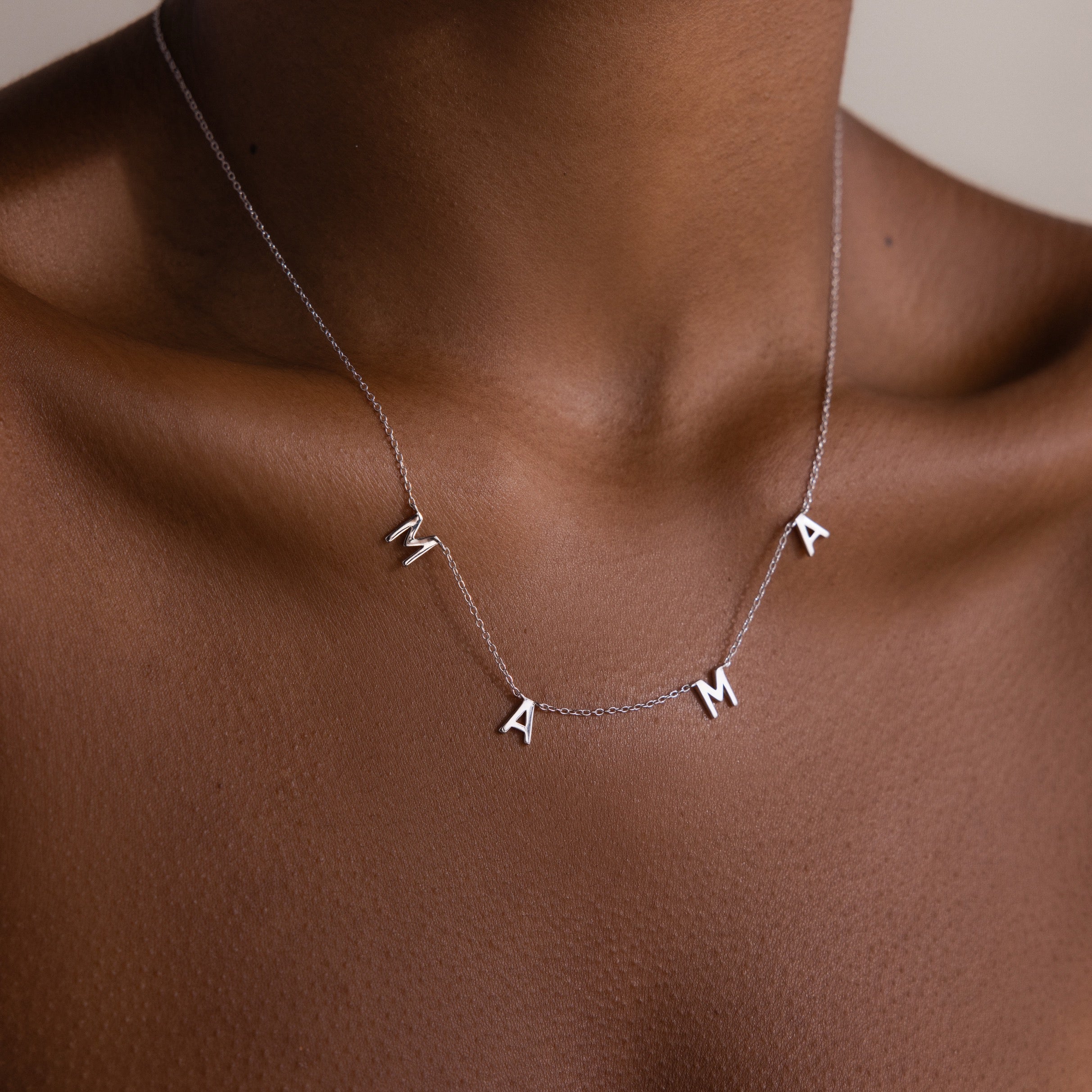The Mama Letter Necklace in Silver spells MAMA and rests elegantly on the neck and collarbone, celebrating motherhood against a neutral background.