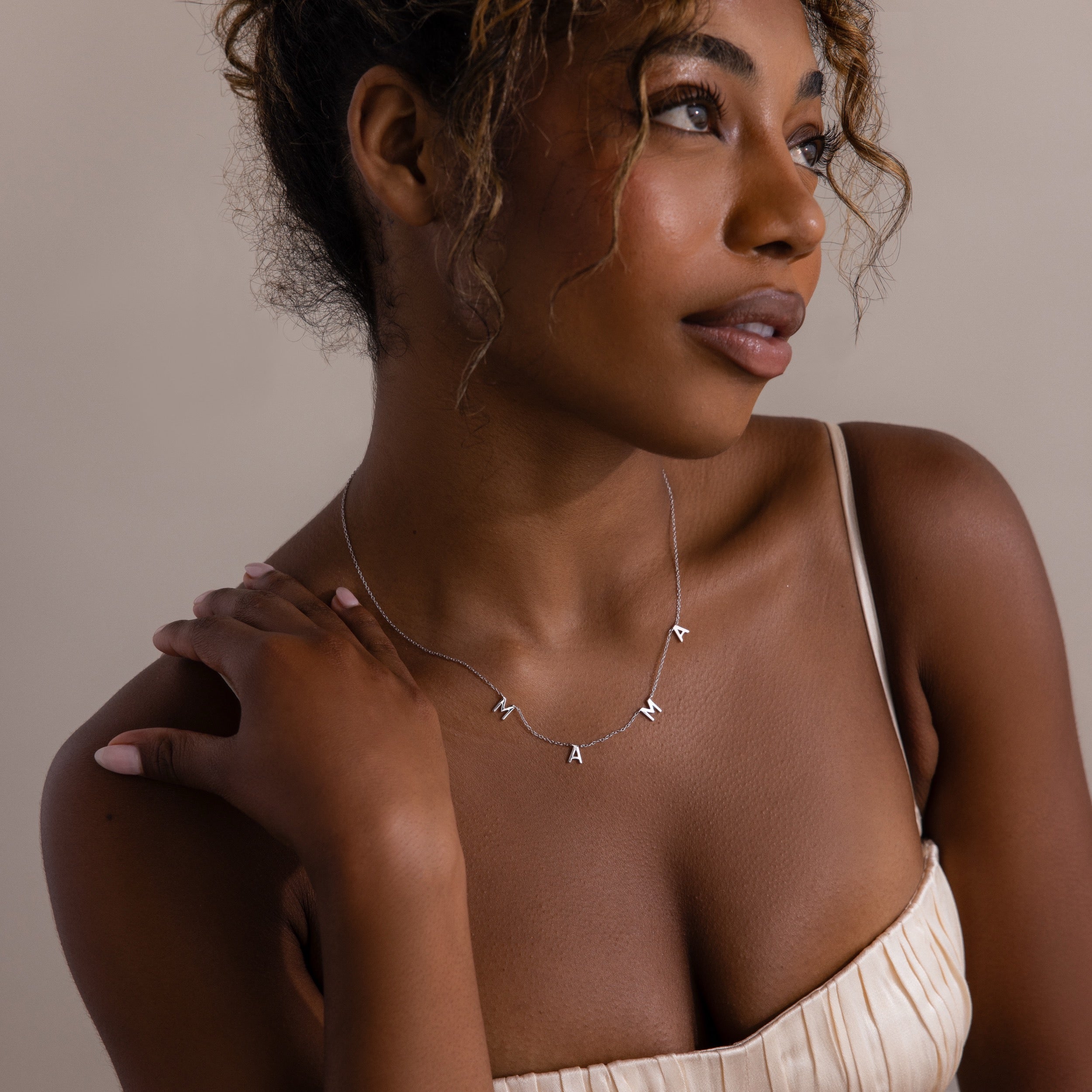 Woman in a cream dress wearing the Mama Letter Necklace in Silver, gazing thoughtfully to the side—a beautiful tribute to motherhood and an ideal gift for mom.