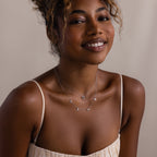 Smiling woman in a cream dress wears layered necklaces, featuring the Mama Letter Necklace in Silver and a heart pendant—a lovely tribute to motherhood.