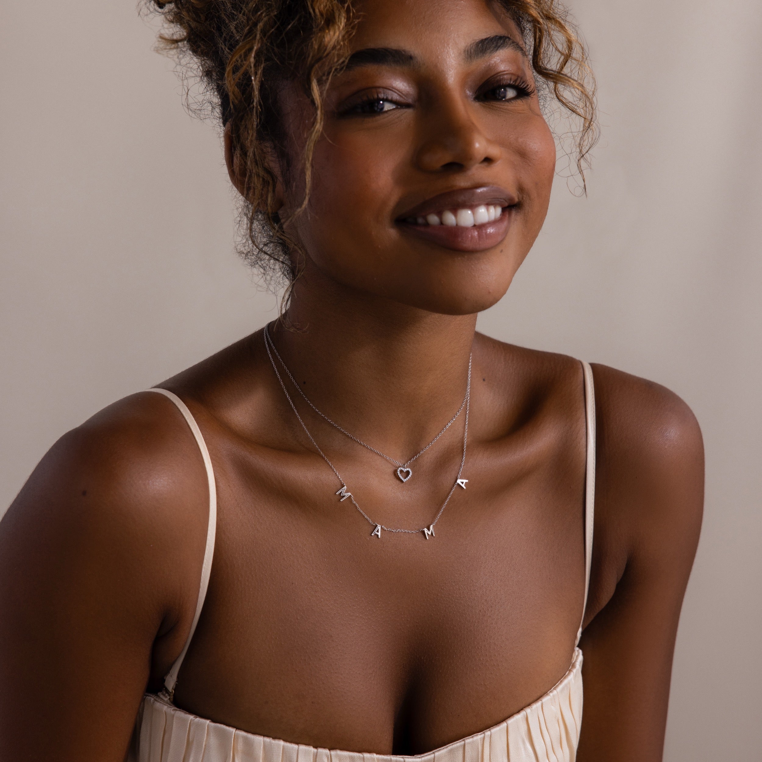 Smiling woman in a cream dress wears layered necklaces, featuring the Mama Letter Necklace in Silver and a heart pendant—a lovely tribute to motherhood.