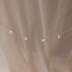 The Mama Letter Necklace in Silver, featuring silver letter charms spelling MAMA, makes a perfect mother gift and is displayed against a soft beige background.
