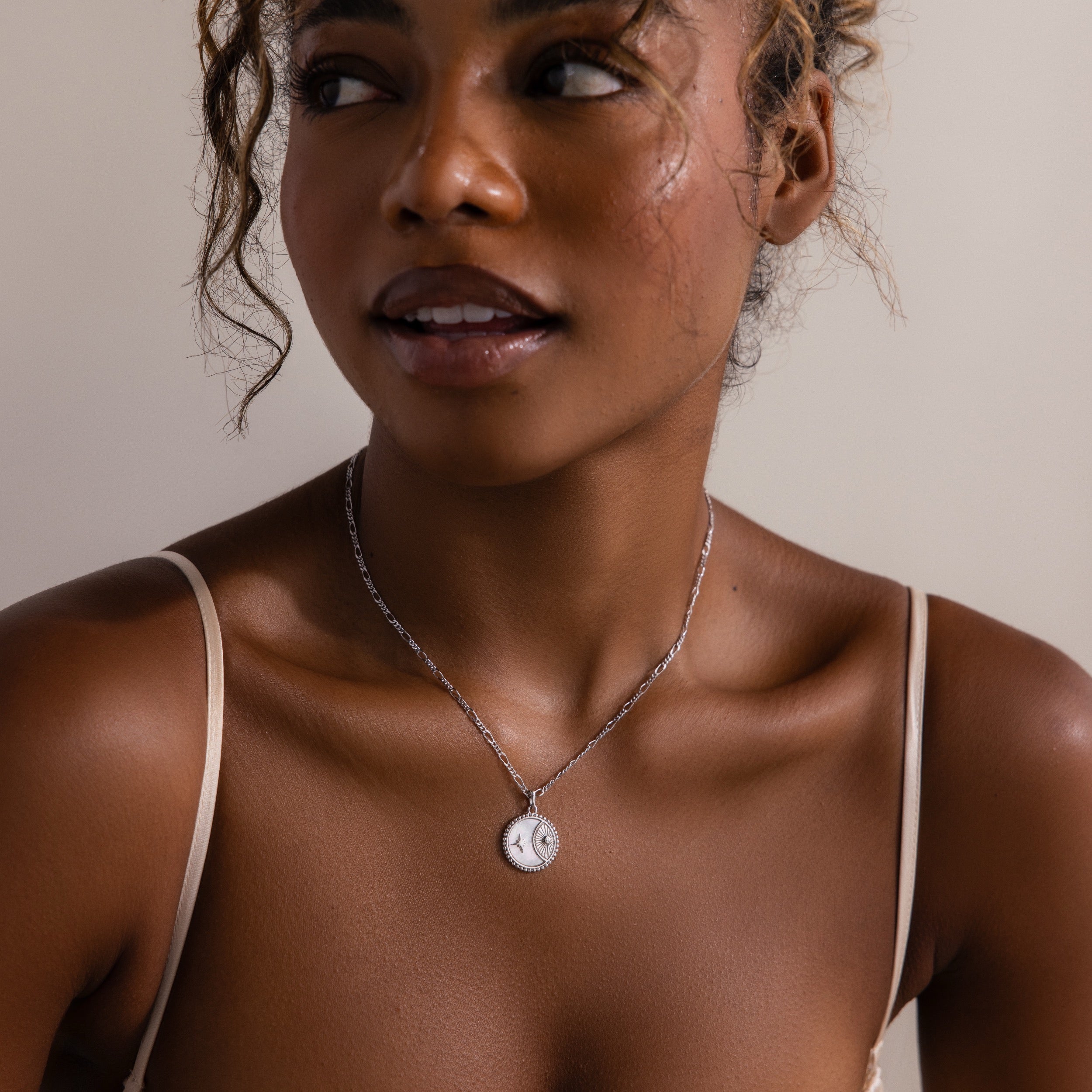 A woman wearing the Gabi Coin Necklace, a minimalist accessory, pairs it elegantly with her cream top while looking to the side and smiling gently.
