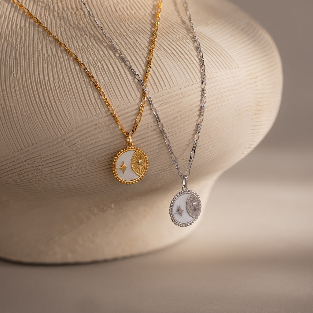 Two Gabi Coin Necklaces, one gold and one silver with round sun and moon pendants, rest on a vase—an ideal minimal set featuring subtle vintage charm.