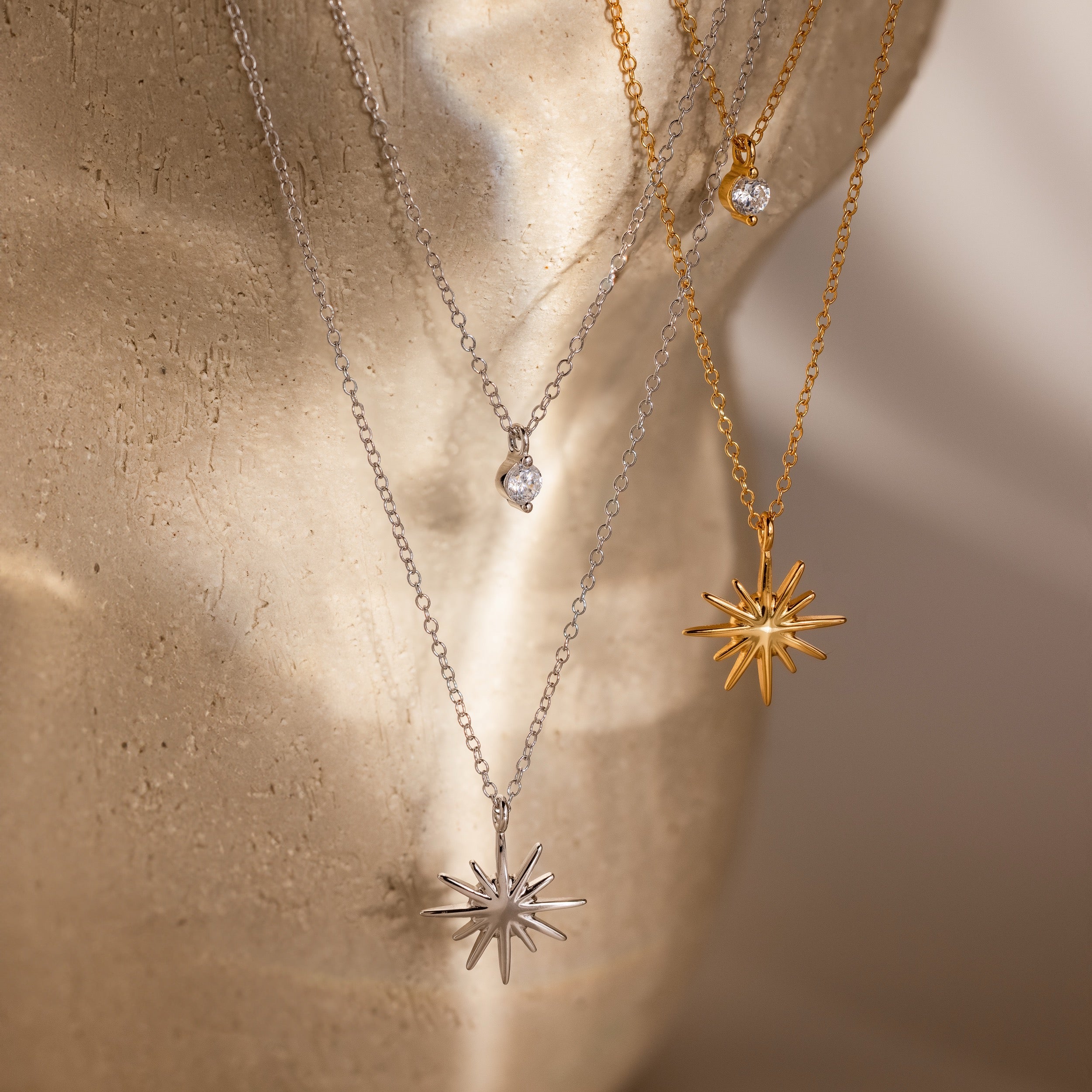 The Laura Starburst Duo Necklaces, featured in gold and silver with starbursts and diamond accents, are displayed on a beige textured surface—ideal for creating a chic layered look.