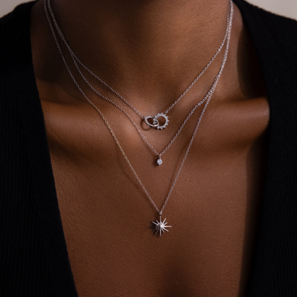 A person wears the Sun  Moon Eternity Necklace, layered with two other silver necklaces featuring unique pendants, against bare skin and a black top.