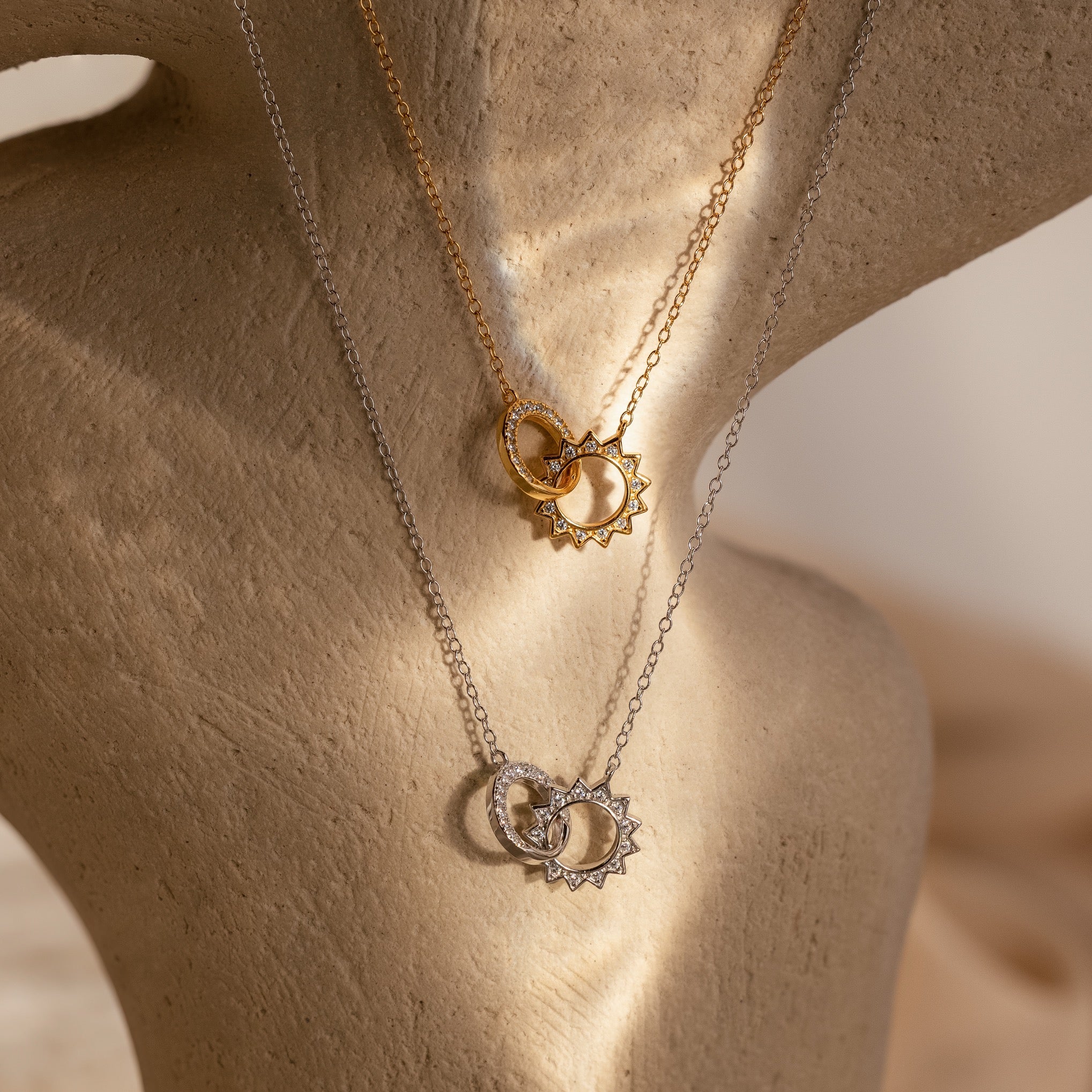 The Sun Moon Eternity Necklaces featured in gold and silver with sun and moon pendants, artfully intertwined and elegantly displayed on a textured stand.