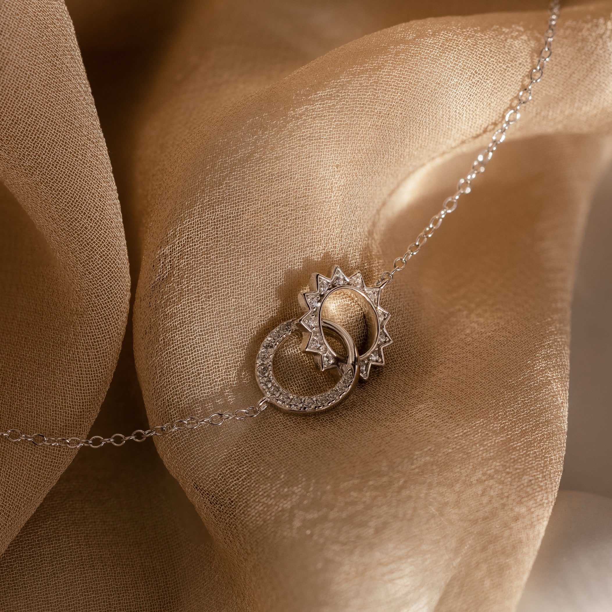 The Sun  Moon Eternity Necklace in Silver features a celestial pendant on a delicate chain, displayed on soft, shimmery beige fabric.